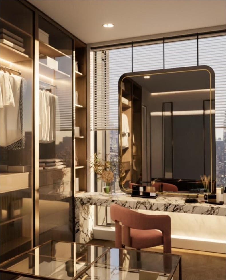 Walk-in dressing room with bronze-framed glass wardrobes, a white marble vanity, a rounded mirror and a rust bouclé chair, lit at dusk.